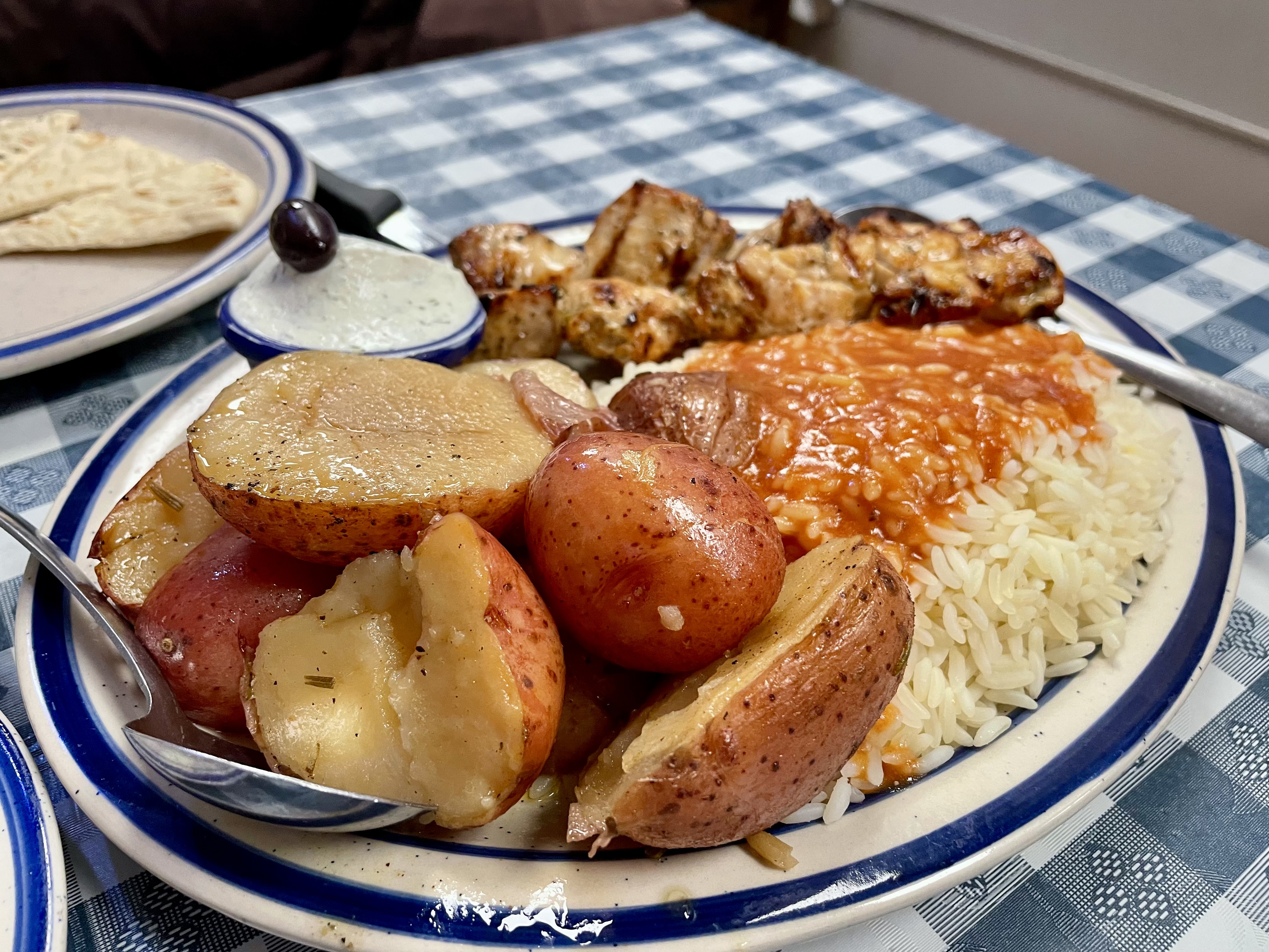 A classic platter featuring tender marinated two souvlaki skewers, roasted potatoes, rice topped with homemade spiro sauce, and a side of Greek salad or soup